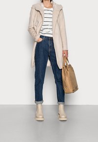 Abrigo beige con capucha sobre una camisa a rayas, combinado con vaqueros oscuros de mezclilla con los puños doblados y botas de tobillo beige. Sostiene un bolso tote color marrón claro.