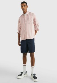 Camicia a righe bianche e rosa con taschino sul petto, abbinata a pantaloni corti blu navy, calze bianche con righe rosse e scarpe nere e bianche.