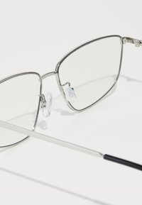 Lunettes de soleil en métal avec des verres rectangulaires. Monture argentée, plaquettes de nez transparentes et accent de tige noir mat élégant. Les verres sont d'un vert clair.