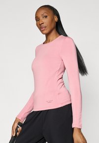 Light pink long-sleeve sport top made of soft fabric, featuring a round neckline and subtle logo detail at the hem. Paired with black pants.