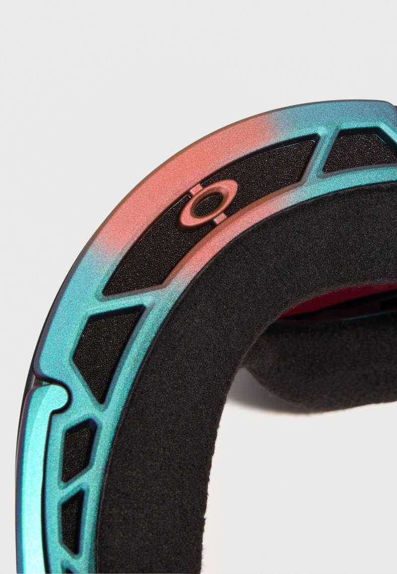 Close-up van een kleurrijke metalen ring met geometrische uitsparingen en zwarte schuimvoering aan de binnenkant, tegen een witte achtergrond.