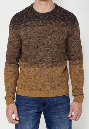 Mann trägt einen langärmeligen Strickpullover mit Farbverlauf in Braun und Senfgelb, kombiniert mit blauen Jeans.