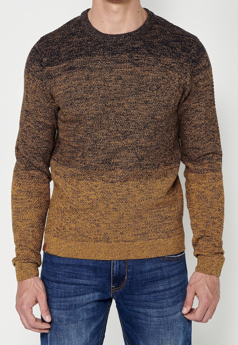Homme portant un pull à manches longues en tricot avec un dégradé de couleurs marron et moutarde, associé à un jean en denim bleu.