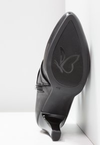 Chaussure à talon noire à bout pointu reposant semelle vers le haut contre un mur blanc, montrant un motif papillon sur la semelle et une fermeture éclair latérale.