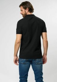 Fekete polo póló rövid ujjal, gallérján díszítő részletekkel és egyenes aljjal. Kék farmernadrággal viselve, texturált anyagból.