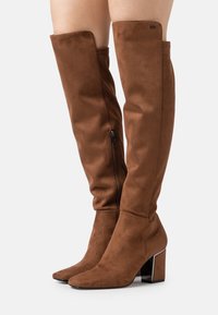 Bottes en daim marron montant au-dessus du genou, avec un bout effilé, une fermeture éclair latérale et un talon métallique épais. Texture lisse avec peu de détails.
