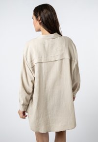 Beige oversized shirt dress made of textured fabric, featuring a back yoke and long sleeves with elastic cuffs.