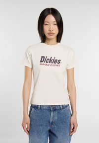 Crème katoenen T-shirt met een voorzijdeafbeelding met "Dickies" en "DUURZAME KLEDING" in donkerrood, gecombineerd met hoge blauwe spijkerbroek.