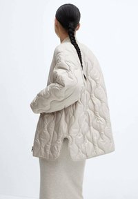 Beige kviltet jakke med en rund, oversized silhuet. Har et tekstureret mønster og en høj-lav kant. Lynlåsdetalje på siden.
