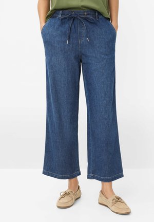 Person iført blå denim bukser med vide ben og snøre i taljen samt beige snørestøvler, stående med hænderne i lommerne.