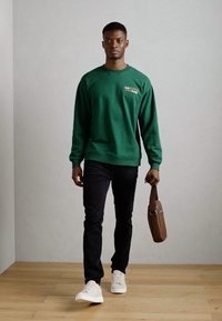 Sweatshirt vert à manches longues, pantalon noir et baskets blanches. Tient un sac marron au design simple. Sol en bois naturel.