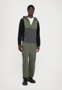 Hoodie com fecho de correr em bloco de cores, em verde-oliva, cinza e preto; combinado com calças de jogging verde-oliva com bolsos cargo e sapatilhas pretas. Textura suave.