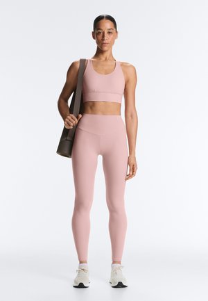 Rózsaszín sportmelltartót és leggingset viselő nő, aki egy feltekert jógaszőnyeget tart a vállán, fehér tornacipőben áll, egyszerű háttér előtt.