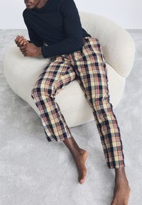 Svart långärmad topp med beiga rutiga byxor med marinblåa och röda accenter. Personen sitter på en texturerad krämfärgad stol.