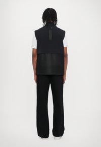 Black sleeveless vest with textured mesh lower panel, featuring a central back loop. Paired with loose black pants and white shirt.