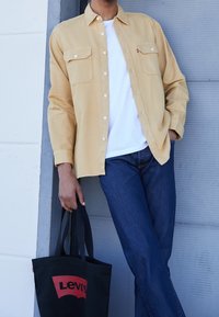 Chemise beige boutonnée avec deux poches poitrine, associée à un jean bleu. Sac fourre-tout noir avec le logo rouge "Levi's". Texture de tissu lisse.