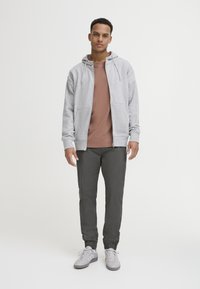 Grey zip-up hoodie with front pockets over a brown t-shirt; paired with dark gray tapered pants and light gray sneakers.