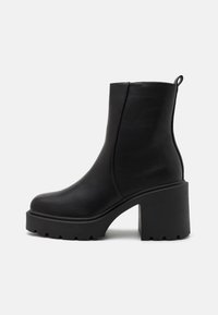 Botines de cuero negro hasta el tobillo con un tacón grueso, textura suave, suela de zigzag y una lengüeta en el talón para facilitar su uso.