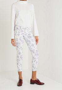 Vit långärmad topp i kombination med blommönstrade slim-fit byxor i vitt och grått. Modellen bär glänsande burgundy skor med snören.