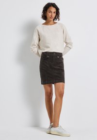 Kvinna iklädd en beige långärmad topp, svart kordflor mini kjol med fickor, och vita sneakers, stående mot en helt vit bakgrund.