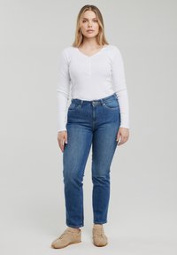 Jeune femme debout, portant un haut blanc à manches longues, un jean bleu et des baskets beige sur un fond gris uni.