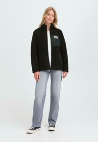 Black fleece jacket with a high collar, zippered chest pocket, and front zipper. Paired with light jeans and black sneakers.