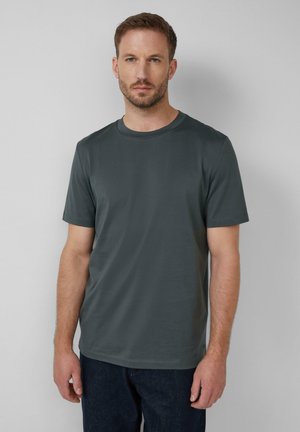 Hombre con cabello corto y barba, vistiendo una camiseta de manga corta gris oscuro y pantalones oscuros, de pie contra una pared gris claro.
