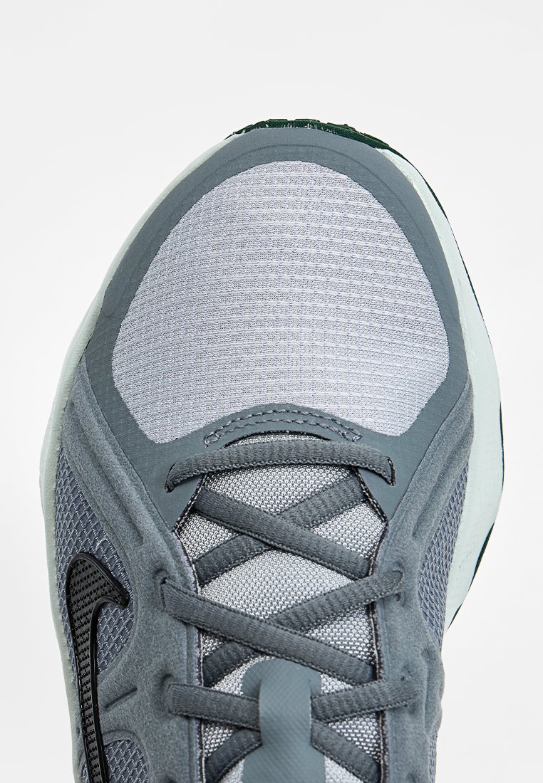 Zapatilla deportiva en malla gris claro con superposiciones en gris más oscuro. Presenta una puntera texturizada con un swoosh negro y cordones planos. Diseño ligero.