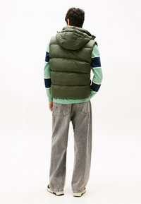 Chaleco acolchado verde oliva con capucha, con secciones acolchadas, llevado sobre una camiseta de manga larga a rayas verdes y azul marino y jeans grises holgados.