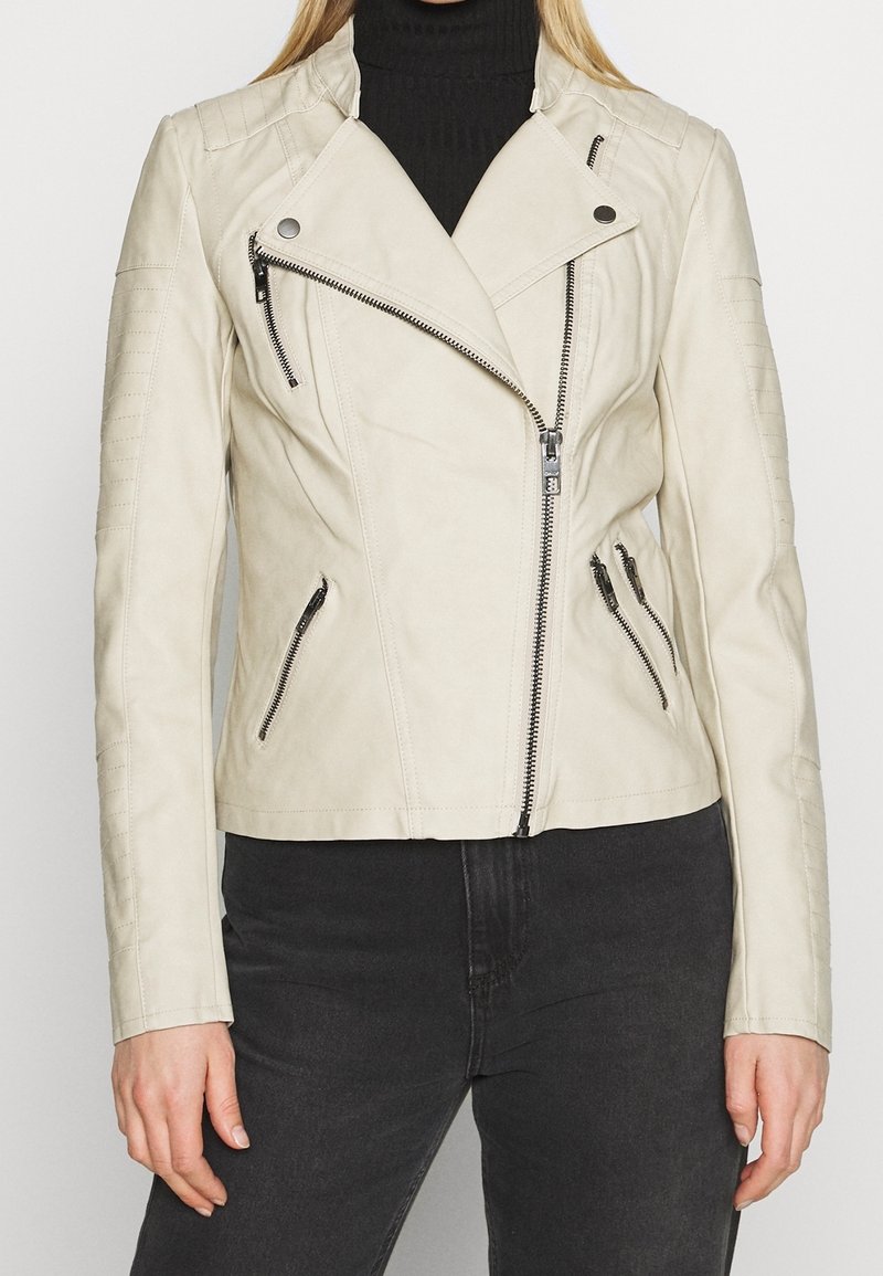 Kobieta ubrana w beżową krótka skórzaną kurtkę motocyklową z asymetrycznym zamkiem błyskawicznym i kieszeniami na zamki, w połączeniu z czarnym golfem i ciemnymi dżinsami.