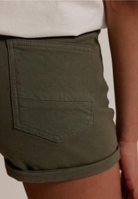 Shorts de algodón verde oliva con dobladillo enrollado, que presentan un bolsillo trasero con un patrón de costura triangular y detalles de costura visibles.