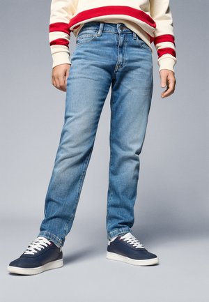 Barn iført lyseblå jeans, marineblå sneakers med hvide såler og en cremefarvet sweater med røde striber på ærmer og kant.