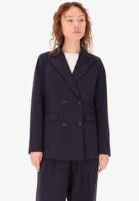 Blazer doppiopetto blu navy in tessuto liscio, caratterizzato da grandi bottoni, revers a scatto e due tasche anteriori. Vestibilità su misura, design minimale.