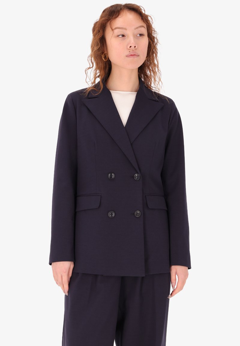 Blazer doppiopetto blu navy in tessuto liscio, caratterizzato da grandi bottoni, revers a scatto e due tasche anteriori. Vestibilità su misura, design minimale.