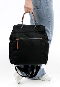 Hand holding black tote bag with front zipper pocket, brown handles, worn with blue jeans and white sneakers on white background.