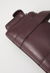 Borsa in pelle bordeaux con una texture liscia, dotata di chiusura a patta e del nome del marchio "Calvin Klein" impresso in argento.