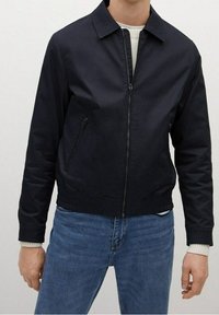 Mann mit einer schwarzen Zip-Jacke mit seitlichen Reißverschlusstaschen über einem weißen Sweater und blauen Jeans, der vor einem einfarbigen Hintergrund steht.