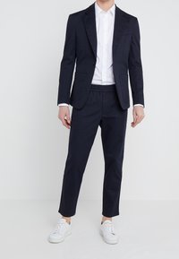 Navy blazer o dopasowanym kroju z wysmukłymi klapami i dwoma przednimi kieszeniami, w połączeniu z granatowymi spodniami elastycznymi w pasie i białymi sneakersami. Gładka tekstura materiału.