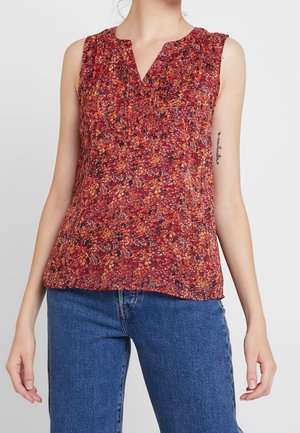Mouwloze rode bloemenblouse met geplooide voorkant, gedragen met blauwe spijkerbroek door een staand persoon tegen een effen achtergrond.