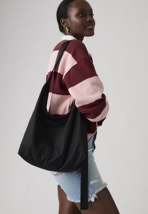 Γυναίκα που φοράει χρυσούς κρίκους στα αυτιά, ριγέ πουλόβερ σε μπορντό και ροζ αποχρώσεις, τζιν σορτς, και κρατάει μια μεγάλη μαύρη τσάντα ώμου σε έναν ώμο.