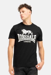 Juodos medvilnės marškinėliai su baltu tekstu "Lonsdale London" ir liūto grafika. Trumpomis rankovėmis, apvalia kaklo iškirpte ir standartiniu pritaikymu.
