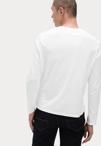 Weißes Langarm-T-Shirt mit rundem Halsausschnitt, glatter Textur und lockerer Passform. Kombiniert mit dunklen Jeans, Ansicht von hinten.