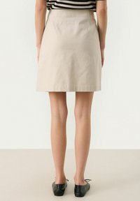 Jupe en coton beige au design évasé et dotée de poches arrière, associée à des chaussures plates noires. Le tissu semble lisse avec un subtil éclat.