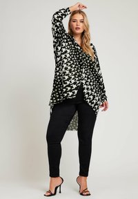 Camisa larga de patrón pata de gallo en blanco y negro, fabricada con tejido ligero, de corte holgado, con dobladillo asimétrico, combinada con pantalones negros ajustados y tacones.