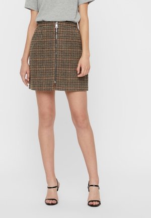 Kobieta ubrana w brązową i czarną mini spódniczkę w kratę z suwakiem z przodu, szary krótki top z krótkim rękawem oraz czarne sandały na wysokim obcasie z paskami.