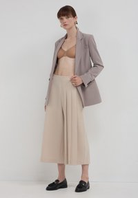 Frau in einem taupefarbenen Blazer, beigen weiten, plissierten Hosen, braunem BH und schwarzen Loafers, die vor einer schlichten hellen Wand steht.