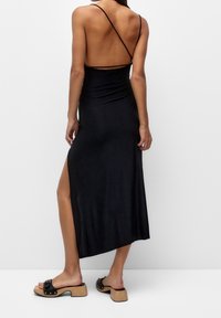 Woman standing facing away wearing a black backless dress with thin straps and a high slit, paired with black heeled sandals.