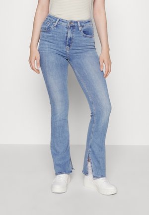 Femme portant un jean taille haute bleu clair avec des fentes à l'avant et des baskets blanches à plateforme, debout devant un fond uni.