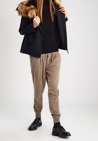 Mustat takki, jossa on tekoturkishtuuli, yhdistettynä beigen värisiin kapeisiin housuihin ja mustiin nauhakengiin. Yksinkertainen muotoilu, matta tekstuuri.