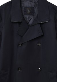 Cappotto doppiopetto blu navy con grandi bottoni neri, chiusura con zip e texture liscia. Presenta una fodera interna a righe e un colletto a revers.